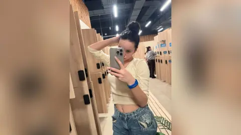 Young woman in a white long-sleeve crop top and denim jeans taking a mirror selfie in a modern clothing store.