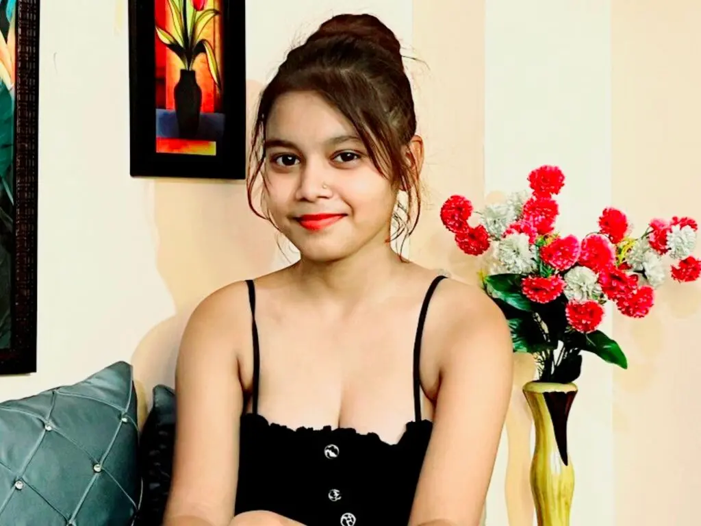 Young woman in a black camisole sitting indoors near red flowers in a bright, relaxed setting.