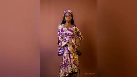 Elegant Black woman in a purple and gold patterned maxi dress sitting against a warm brown studio background.