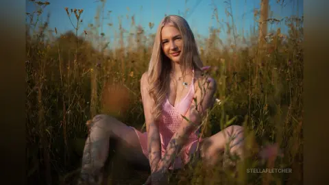 StellaFletcher sits in a field of tall grass wearing a pink mesh bodysuit with a deep neckline.