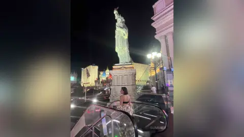 Woman in a patterned top standing by stairs near the illuminated Statue of Liberty replica at night.
