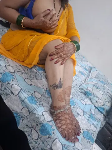 Woman in yellow traditional outfit with intricate henna patterns sitting on a blue floral patterned bedsheet.