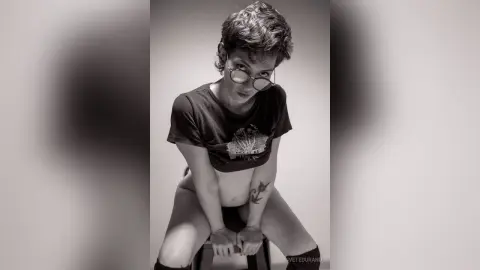 IveteDurand wears glasses and a black t-shirt while sitting and looking down at the camera in this black and white photo.