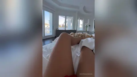 Relaxed woman in white loungewear resting on a bed in a bright, modern sunlit bedroom.