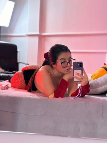 Young woman in red velvet cosplay outfit and glasses taking a selfie on a bed in soft pink lighting.