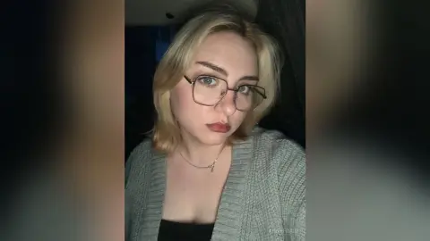 AnnyFraid wears glasses and a grey cardigan over a black top, looking directly at the camera with blonde hair and red lipstick.