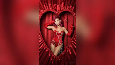 Glamorous woman in a red bodysuit and long gloves posing inside a heart-shaped frame against red velvet curtains.