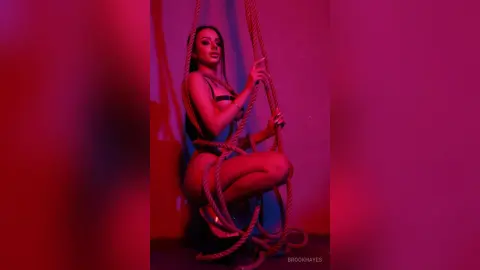 Glamorous woman in black lingerie sitting on ropes under moody pink neon lighting.