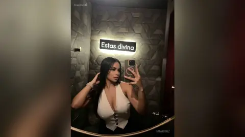 MiaKorz takes a mirror selfie wearing a tight white sleeveless top while posing in front of a textured wall.