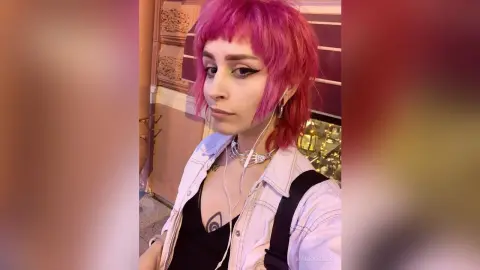 Young woman with pink hair and winged eyeliner wearing a white denim jacket in soft moody lighting.