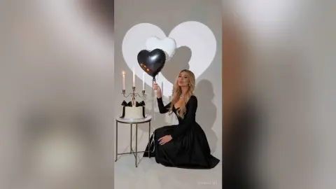 SerenaStar poses in a long black dress next to a white cake and heart-shaped balloons.