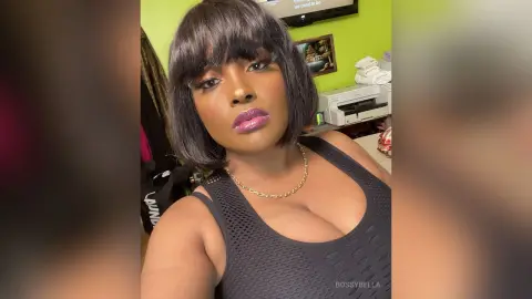 BossyBella — BossyBella poses in a black mesh tank top and gold chain, showing off her dark bob haircut and purple lipstick in a room with lime green walls.