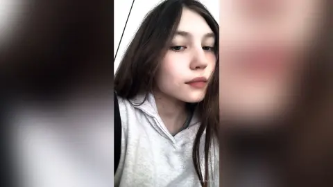 Young woman with long dark hair wearing a cozy grey hoodie in a soft, natural indoor setting.