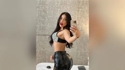 Young woman in a sparkly black crop top and shiny leather pants taking a confident mirror selfie indoors.