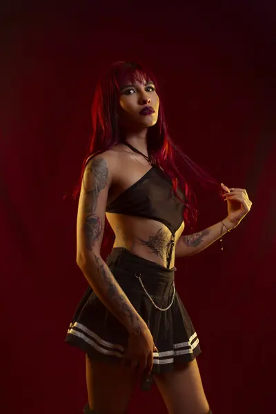 Naila_Mistress poses in a red lace bodysuit, showing her cleavage and legs while sitting on a white chair against a warm, blurred background.