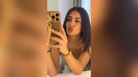 Young woman in a white tank top taking a playful mirror selfie with a gold smartphone.