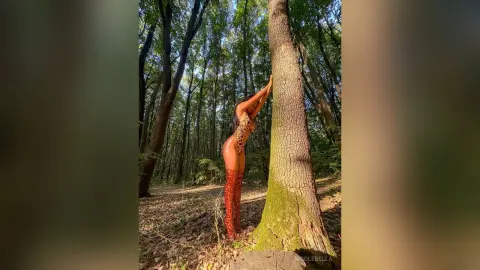 NicoleBella stands naked against a large tree trunk in a sunlit forest, wearing red patterned lace lingerie.