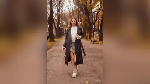 Stylish woman in a black trench coat and white sweater walking through a peaceful autumn park path.