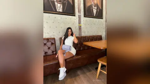 Stylish woman in a white mini dress and white sneakers sitting on a brown leather sofa in a chic cafe.