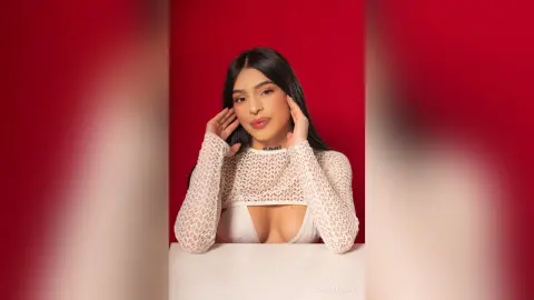 Young woman in a white lace long-sleeve top posing against a red background with a confident mood.