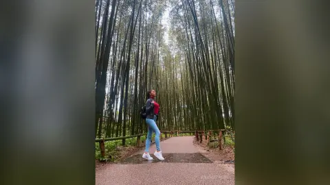 AnaMathers stands on a path surrounded by tall bamboo trees, wearing a red top and blue jeans.