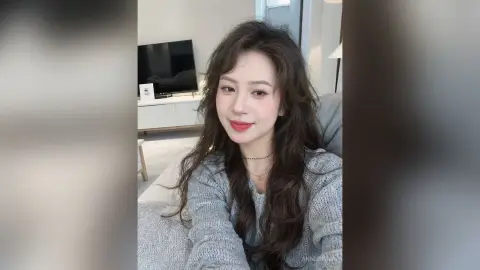 Smiling young woman with long wavy hair wearing a cozy grey sweater in a bright modern living room.