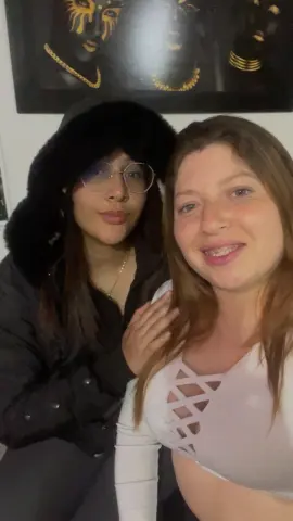 Two smiling young women posing together indoors, one in a black winter coat and fuzzy hat, the other in a white top.