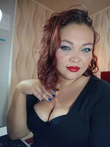 Red-haired woman in a black dress posing with a confident expression in a bright indoor setting.