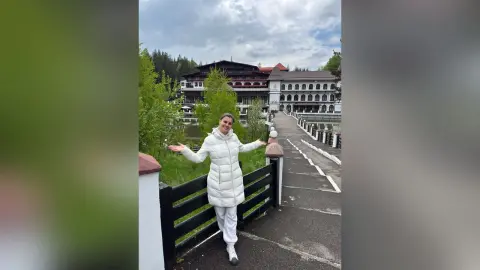 AnishaBee stands outside on a path wearing a white puffer jacket and white pants, smiling with her arms spread wide near a lake and large building.
