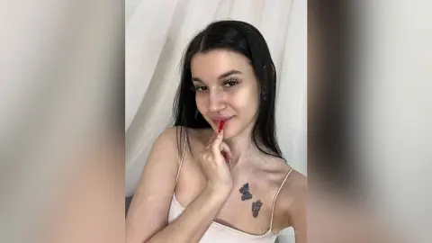 Smiling young woman with long dark hair and butterfly tattoo wearing a white camisole in a bright indoor setting.