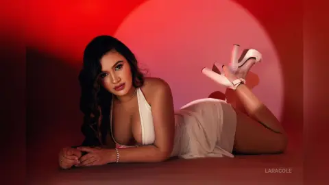 Glamorous woman in a white halter dress and white heels lying on a red studio background.