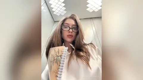 Young woman with long brown hair and glasses holding a notebook in a modern elevator setting.