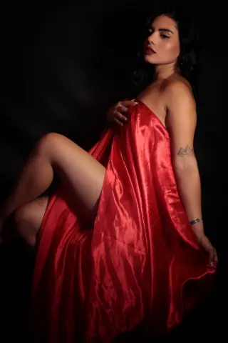 Elegant woman in a flowing red satin gown posing against a dark background for a moody, glamorous portrait.