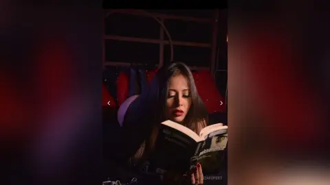 Young woman in a dark outfit reading a book in a moody, dimly lit room.