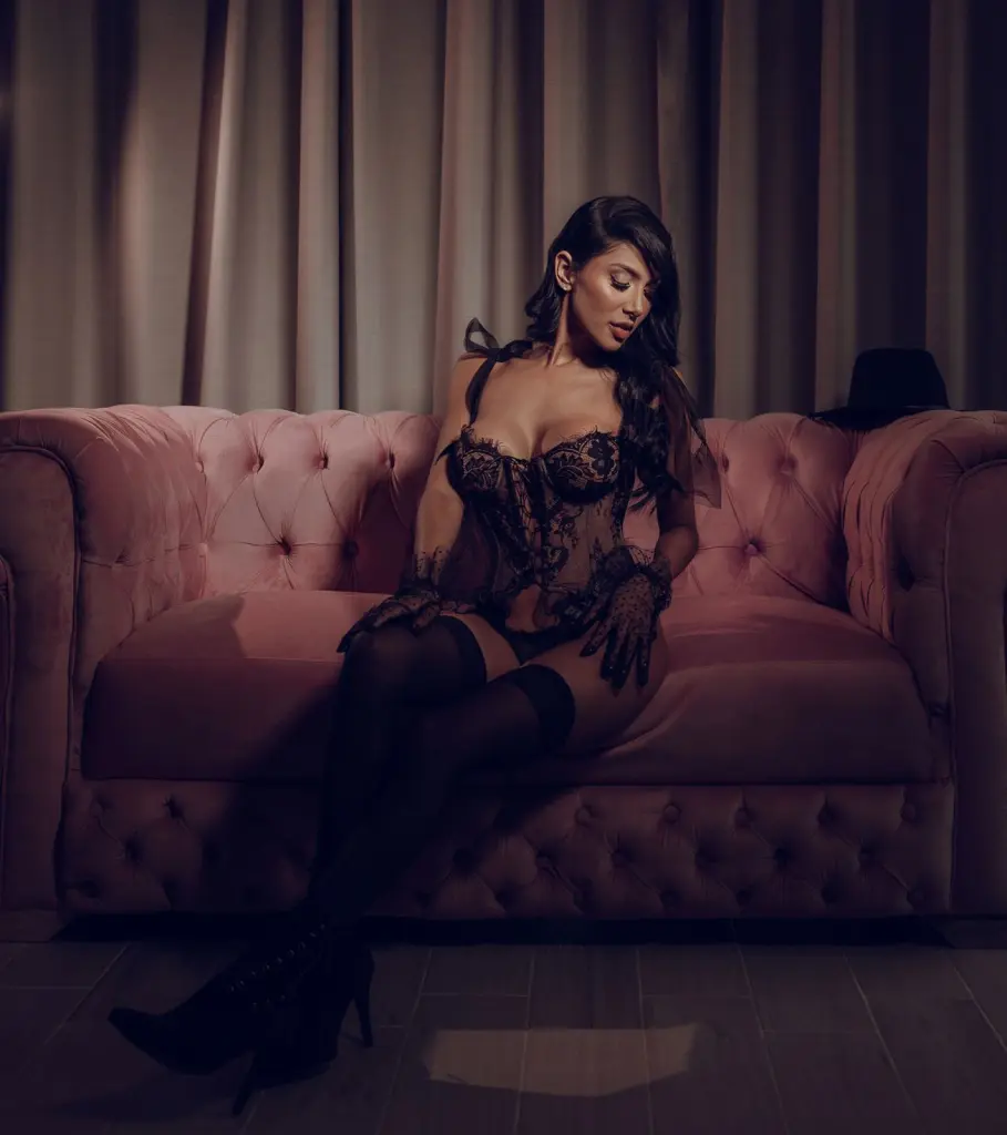 AnnyHoney poses in a black corset top and black pants, showing off her long dark hair and cleavage.