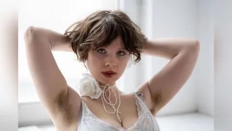 Young woman with wavy hair in a white lace dress posing with hands behind head in a bright, airy room.