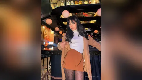 Stylish woman in a white turtleneck and brown skirt posing in a moody, dimly lit cocktail bar.