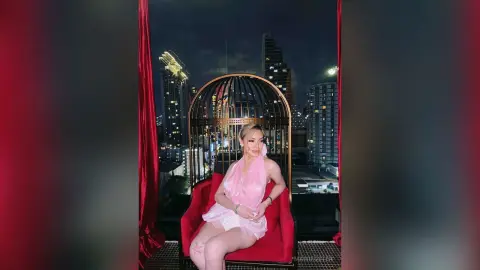 Glamorous woman in a pink outfit sitting in a decorative birdcage chair on a luxury city rooftop at night.