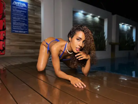 Curvy brunette woman in blue bikini posing on a wooden deck by a luxury pool at night.