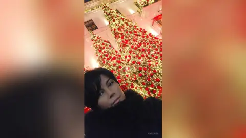 Woman in a black faux fur coat smiling in front of a glowing, festive Christmas tree at night.