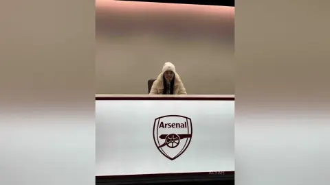 Smiling woman in a white winter coat sitting behind an Arsenal FC branded desk in a modern office setting.