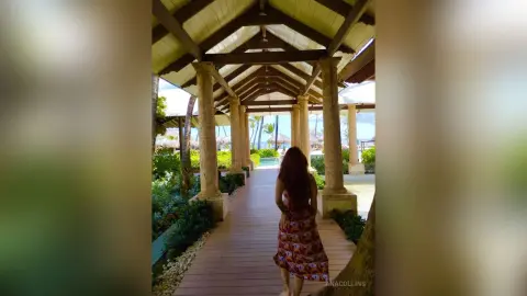Woman in a floral summer dress walking through a tropical wooden pavilion toward a sunny beach view.