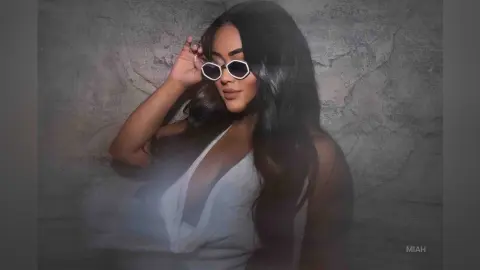 Glamorous woman in white plunging dress and white sunglasses posing against a textured stone wall.
