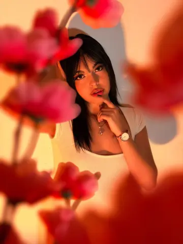 DapneOwen poses with long dark hair and red lipstick, framed by blurry pink flowers in warm lighting.