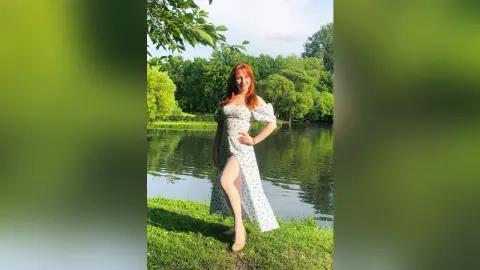 Redheaded woman in a white floral sundress standing by a serene lake in a lush green park.