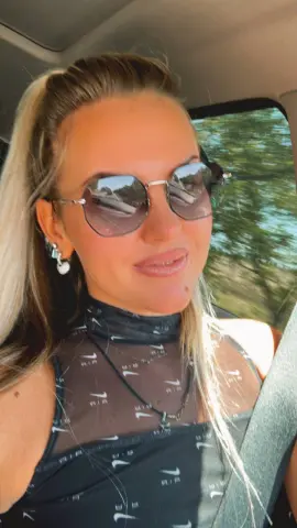 Smiling blonde woman in a black mesh top and sunglasses sitting in a car on a sunny day.