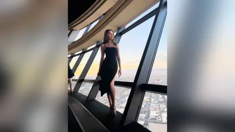 EvelynMills stands on a high balcony wearing a black sleeveless dress with a high slit, looking out over a city view.