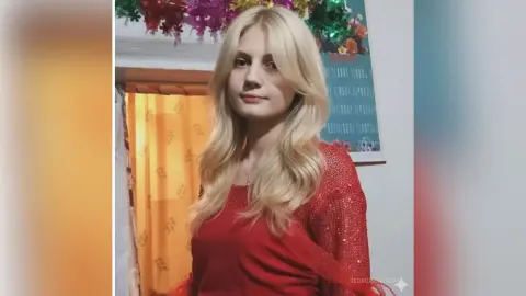 GeorgineAragan poses in a red sequined top with long blonde hair against a background of yellow curtains and a wall poster.
