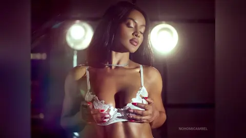 NohomiCambel poses in white lace lingerie, holding her bra or bra cups while looking down under bright studio lights.