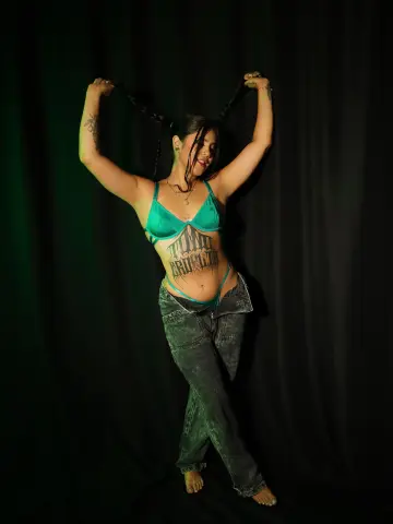 Tattooed woman in a teal bikini top and denim jeans posing playfully with long braids against a dark backdrop.